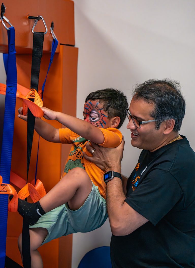 Child with face paint climbing on harness with adult support in pediatric therapy setting, emphasizing independence and skill development.